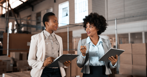 Two professional women discussing work while holding clipboards and a tablet in a warehouse with stacked boxes.