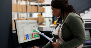 Female warehouse worker reviewing inventory on a computer screen while holding a clipboard, with shelves of boxes in the background.